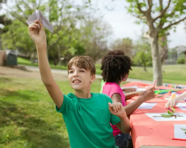 Chłopiec puszcza papierowego samolocika, zastanawiając się, gdzie wysłać dziecko na obóz językowy. Dzieci tworzą prace plastyczne na stole w parku.