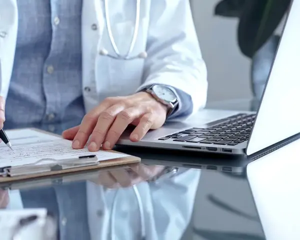 Lekarz w białym kitlu pisze na dokumentach, obok laptop. Szybka konsultacja psychiatryczna online.