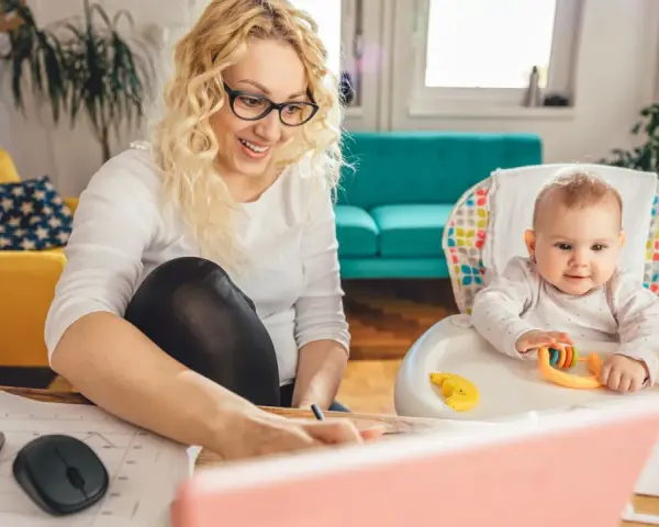 Uśmiechnięta mama pracuje przy laptopie, podczas gdy dziecko bawi się w krzesełku. Planuje zwrot podatku na dziecko.