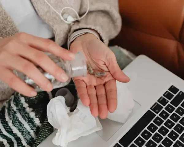 Dłoń z kapsułkami, obok laptop i chusteczki. Kto płaci za L4, gdy choroba dopada?
