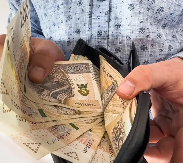 Dłonie trzymają portfel z banknotami 200 zł. Ile trzeba przepracować, by tyle zarobić?