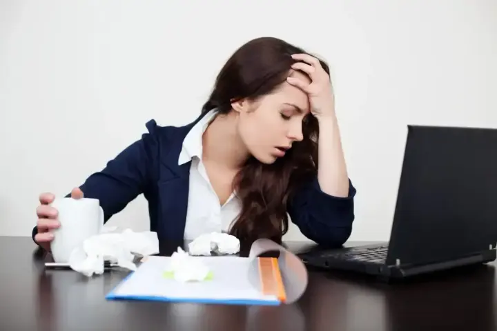 Zmęczona kobieta z kubkiem i chusteczkami przy laptopie. Zastanawia się, ile można być na zwolnieniu lekarskim od psychiatry.