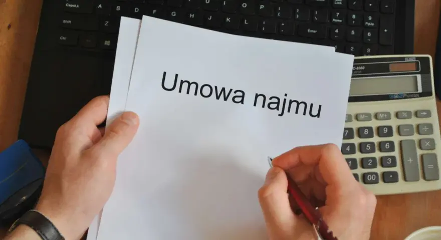 Ręka pisze długopisem na umowie najmu, obok kalkulator i laptop. Czas pomyśleć o podatku od najmu.