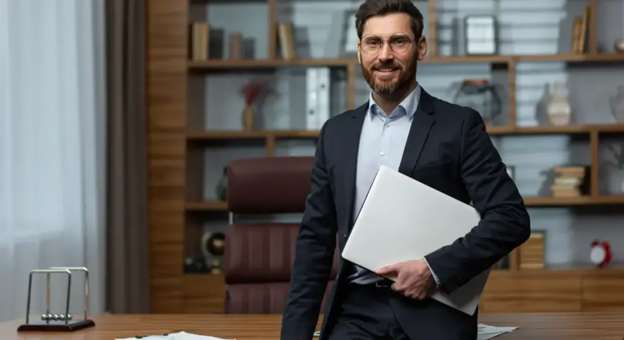 Uśmiechnięty dyrektor szkoły w garniturze, trzymający laptopa, stoi przy biurku w gabinecie.