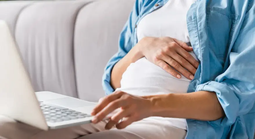Kobieta w ciąży siedzi na kanapie, pisząc na laptopie wniosek o urlop macierzyński.
