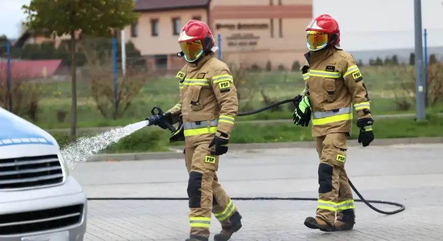 Dwóch strażaków w akcji. Jeden z nich polewa wodą samochód, drugi niesie wąż. Ciekawe, ile zarabia strażak za taką pracę.