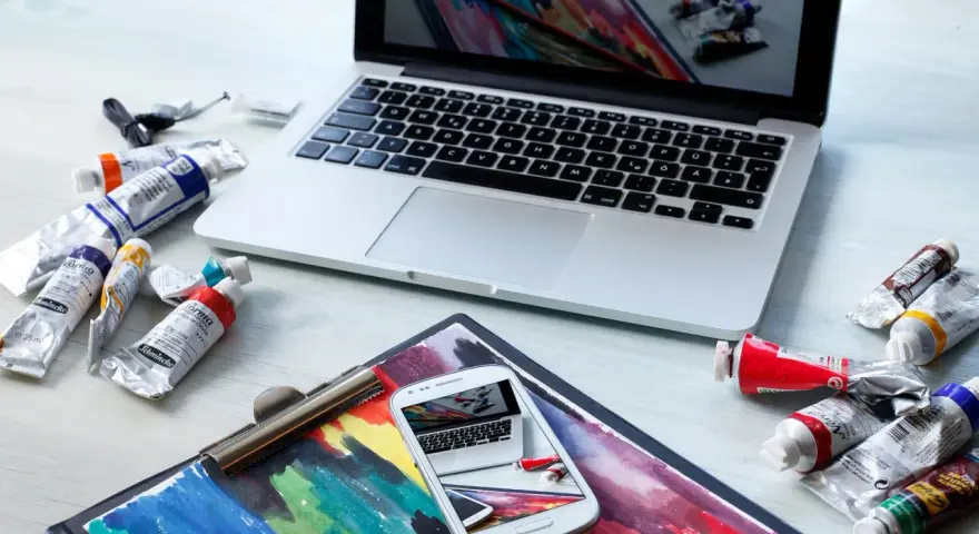 Na biurku leżą farby i laptop, na ekranie którego widać podobny obraz. Zastanawiasz się, ile zarabia grafik komputerowy?