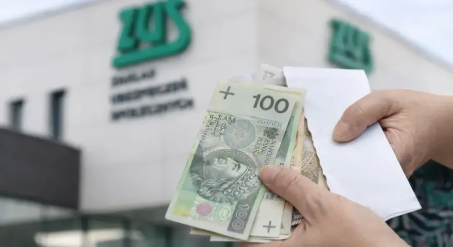 Ręce trzymają polskie banknoty przed budynkiem ZUS. To symboliczny moment, kiedy wypłata 14 emerytury staje się rzeczywistością.