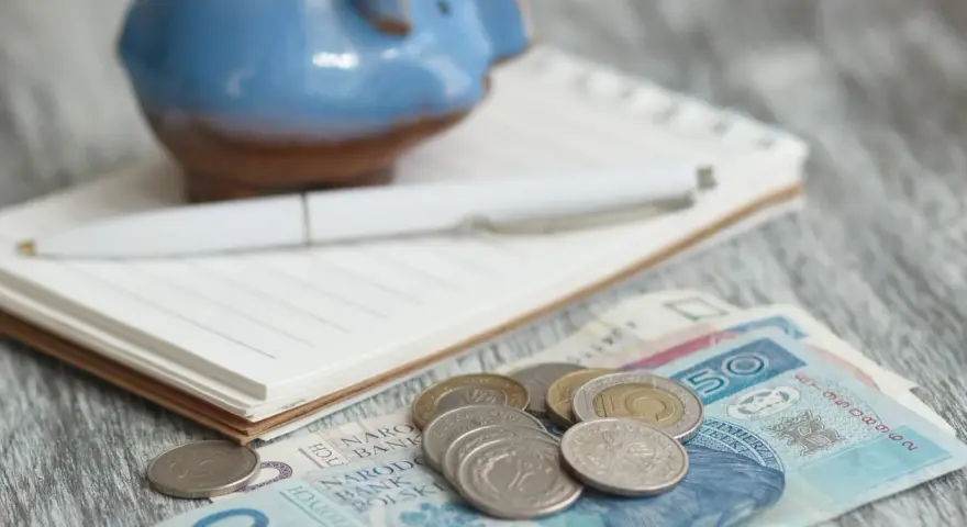 Świnka skarbonka, notatnik i długopis nad polskimi banknotami i monetami. Zastanawiasz się, ile wyniesie waloryzacja kapitału początkowego w 2025?