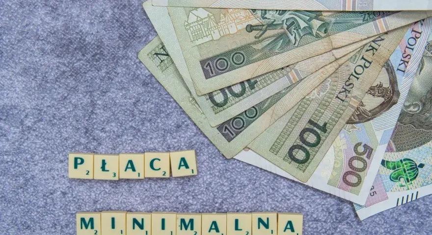 Płaca minimalna w Polsce – polskie banknoty o nominałach 100 i 500 zł ułożone na szarym tle, obok kafelki z literami tworzące napis "PŁACA MINIMALNA".