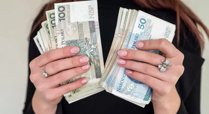 Dłonie z paznokciami w kolorze nude trzymają stosy banknotów 100 i 50 zł. To idealna jubileuszówka na prezent!