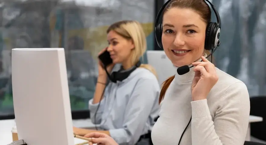 Uśmiechnięta key account manager w słuchawkach z mikrofonem pracuje przy komputerze. W tle koleżanka rozmawia przez telefon.