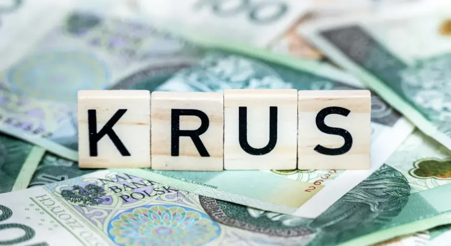 Drewniane klocki z napisem "KRUS" leżą na stosie polskich banknotów stuzłotowych. To symbol oszczędności na emeryturę z KRUS.