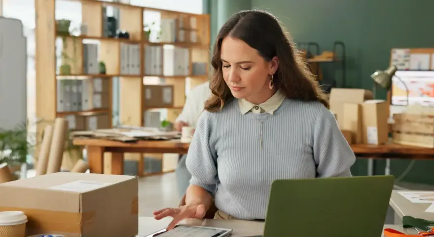 Kobieta pracuje przy biurku, na którym leży tablet z danymi. W tle widać regały z paczkami. Może to być związane z ksef odroczenie.