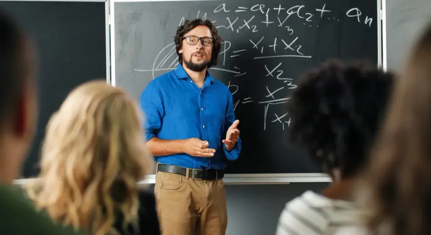 Nauczyciel w niebieskiej koszuli tłumaczy matematykę przy tablicy. Czy nauczyciel jest funkcjonariuszem publicznym? To pytanie nurtuje wielu.