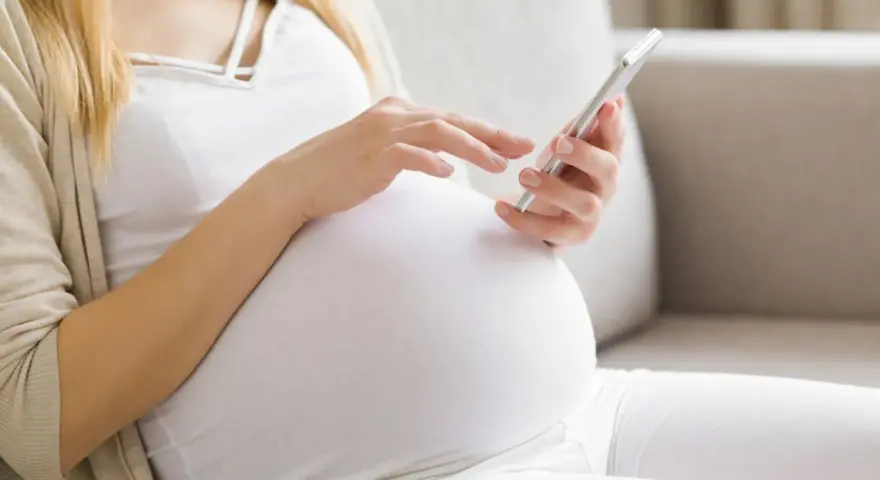 Kobieta w 34. tygodniu ciąży przegląda coś na telefonie, siedząc na kanapie.