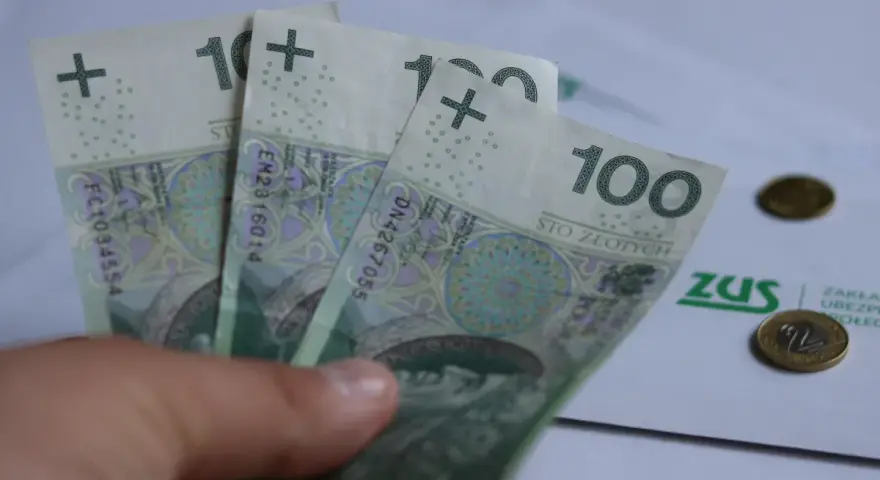 Trzy stuzłotowe banknoty w dłoni, obok dokument ZUS i monety. Czy to zapowiedź 30 krotności ZUS 2025?