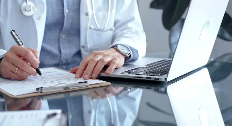 Lekarz w białym kitlu pisze na dokumentach, obok laptop. Szybka konsultacja psychiatryczna online.