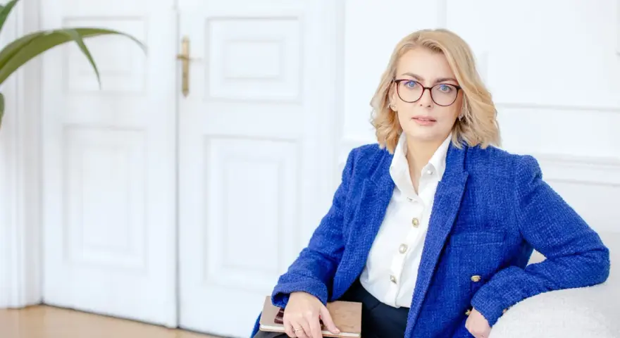 Kobieta w okularach i niebieskiej marynarce trzyma notatnik. Planuje dofinansowanie na otwarcie firmy.