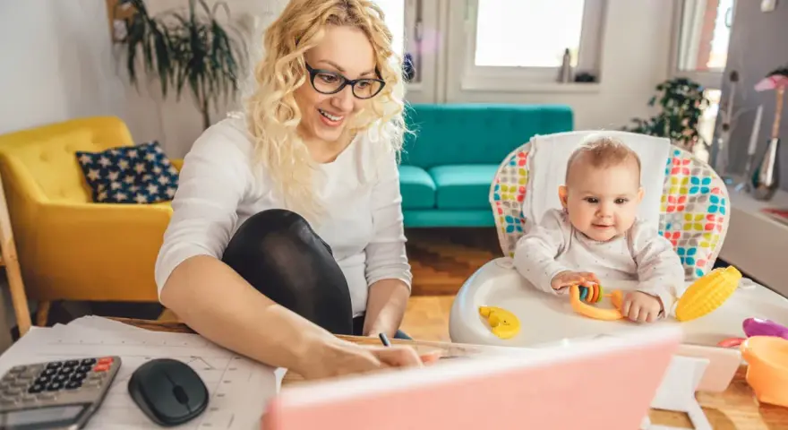 Uśmiechnięta mama pracuje przy laptopie, podczas gdy dziecko bawi się w krzesełku. Planuje zwrot podatku na dziecko.
