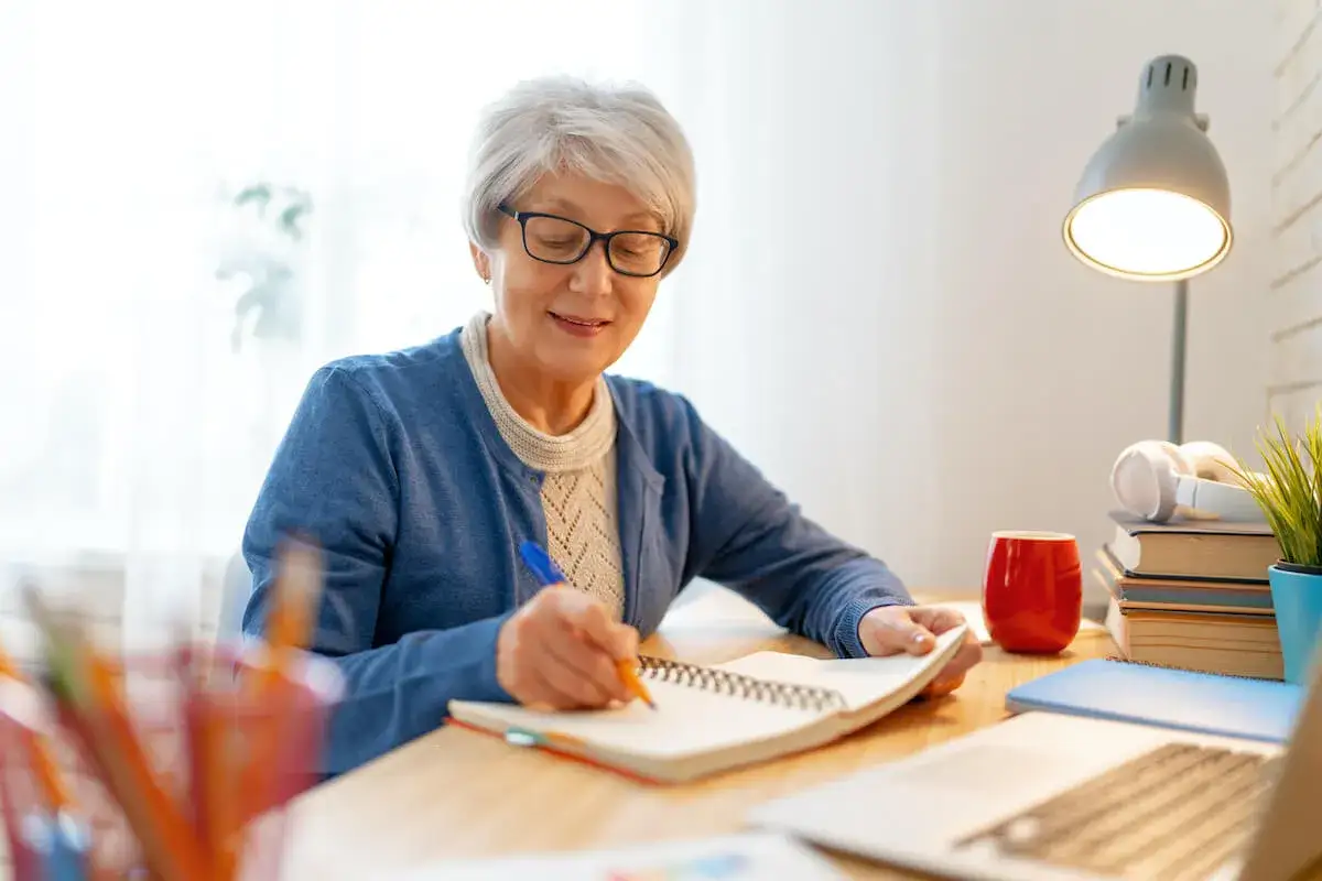 Uśmiechnięta seniorka pisze w notatniku, ciesząc się ulgą dla pracujących seniorów. Na biurku laptop, lampka i kubek.