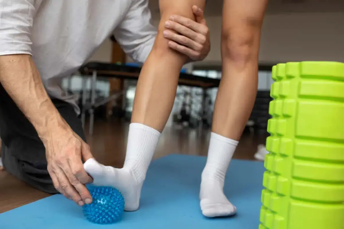 Fizjoterapeuta masuje stopę pacjenta z użyciem piłeczki, pomagając w rehabilitacji po skręceniu stawu skokowego. Pytanie o ile L4 na skręcenie stawu skokowego jest tu nie na miejscu.