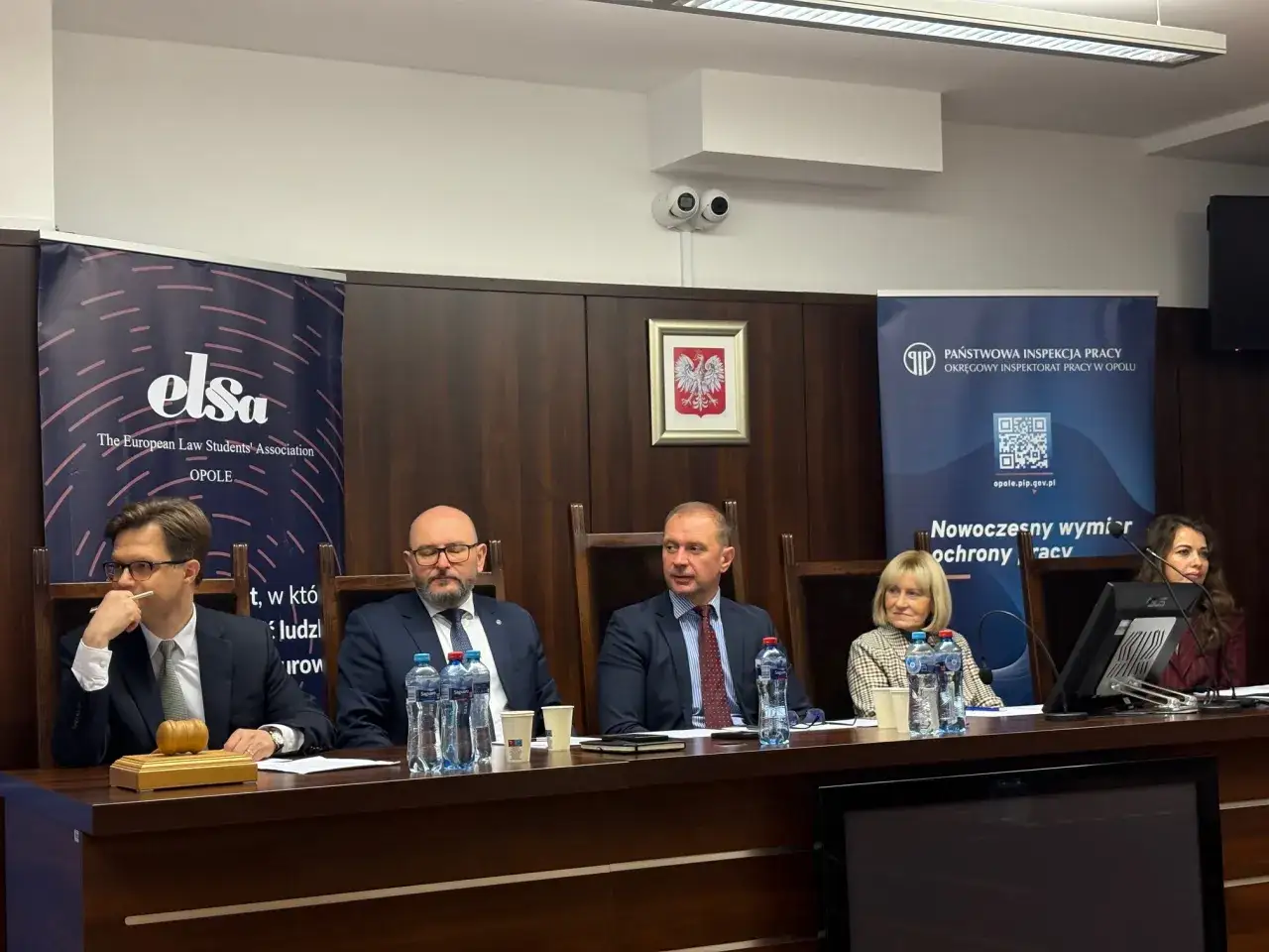 Panel dyskusyjny z udziałem studentów prawa. Na stole młotek sędziowski i banery ELSA oraz Państwowej Inspekcji Pracy.
