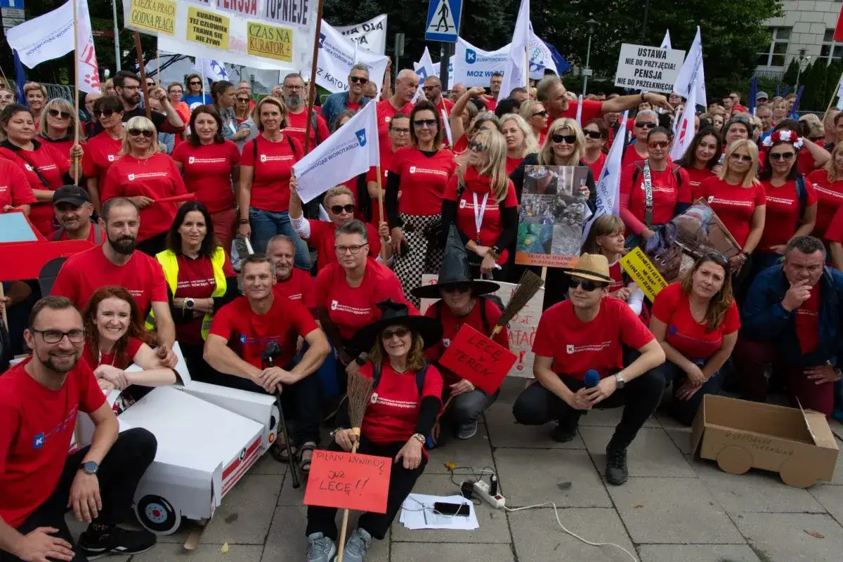 Protest kuratorów sądowych w czerwonych koszulkach. Trzymają transparenty z hasłami o godnej płacy i lepszych warunkach pracy.