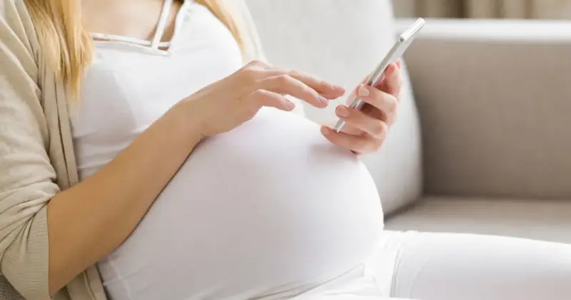 Kobieta w 34. tygodniu ciąży przegląda coś na telefonie, siedząc na kanapie.