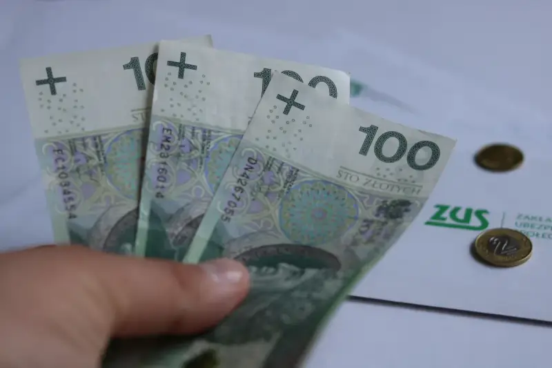 Trzy stuzłotowe banknoty w dłoni, obok dokument ZUS i monety. Czy to zapowiedź 30 krotności ZUS 2025?